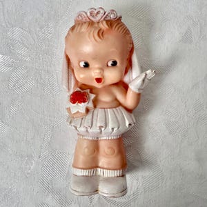 May include: Vintage bisque figurine of a baby bride. The figurine has a pinkish skin tone, a white skirt, and white gloves. The baby is wearing a crown and holding a red flower bouquet. The figurine is standing on white boots.