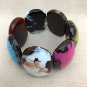 Könnte beinhalten: Ein farbenfrohes Armband aus runden, flachen Glasperlen. Die Perlen sind braun, schwarz, weiß, blau, rosa und gelb mit wirbelnden Mustern.