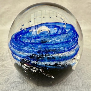May include: A clear glass paperweight with a swirling blue and white design. The bottom of the paperweight is black. Small air bubbles are visible throughout the glass, adding to its decorative appeal.