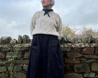May include: A woman wearing a cream-colored cable-knit sweater, a navy blue skirt with pockets, and white sneakers with black stripes. She is standing in front of a stone wall, looking up at the sky. The sweater has a small red detail.
