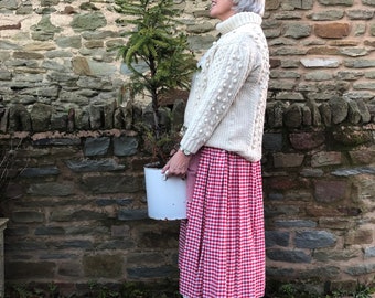 May include: A woman wearing a white cable knit sweater with a red and white gingham skirt. She is holding a white pot with a small evergreen tree.