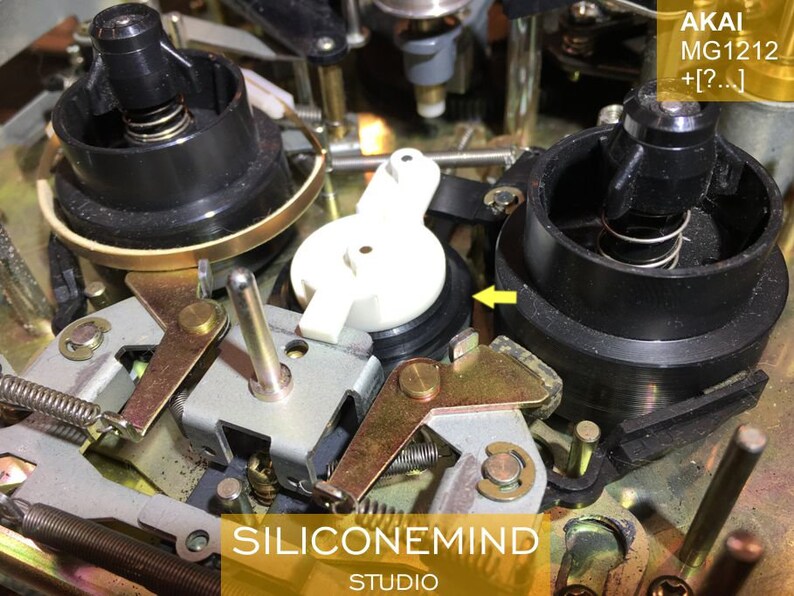 May include: Close-up of the inner workings of an Akai MG1212 cassette deck. The image shows a black plastic reel with a spring, a white plastic gear, and a gold metal lever.