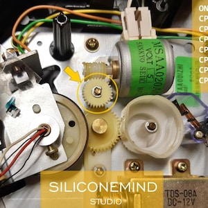 May include: Close-up of a small, white plastic gear with a gold metal center. The gear is part of a larger mechanism, likely from a cassette player or other electronic device. The gear is surrounded by other gears and wires.