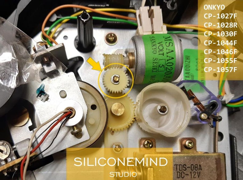 May include: Close-up of a small, white plastic gear with a gold metal center. The gear is part of a larger mechanism, likely from a cassette player or other electronic device. The gear is surrounded by other gears and wires.