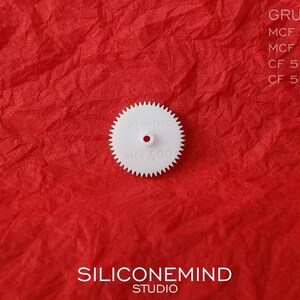 Puede incluir: Engranaje de pl&aacute;stico blanco con el texto "GRUNDIG MCF 500" impreso en &eacute;l. El engranaje est&aacute; sobre una superficie roja texturizada.