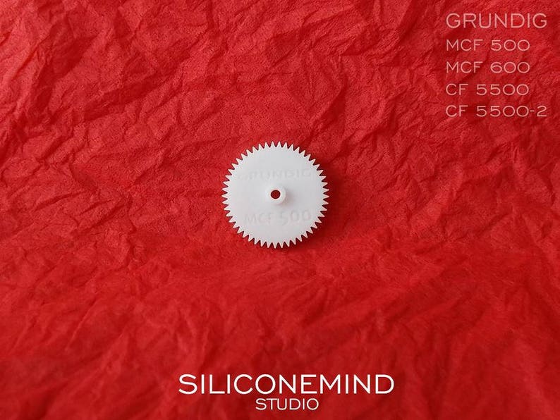 Puede incluir: Engranaje de pl&aacute;stico blanco con el texto "GRUNDIG MCF 500" impreso en &eacute;l. El engranaje est&aacute; sobre una superficie roja texturizada.