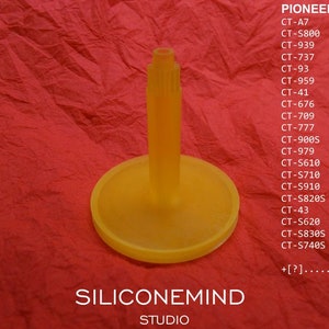 Op de afbeelding: Een gele, cilindrische component met een brede, cirkelvormige basis en een centrale steel. De afbeelding bevat een lijst met Pioneer-modelnummers en de tekst "SILICONEMIND STUDIO". De achtergrond is een getextureerd rood oppervlak.