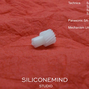 May include: A small, white plastic gear with a cylindrical body and toothed edge, set against a textured red background. The image includes text identifying the gear as a mechanism unit for Panasonic and Technics models.