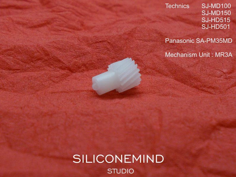 May include: A small, white plastic gear with a cylindrical body and toothed edge, set against a textured red background. The image includes text identifying the gear as a mechanism unit for Panasonic and Technics models.