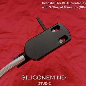 Può includere: Testina nera per giradischi DUAL con bracci a forma di S. La testina ha due fori per viti e un braccio argentato attaccato. Il testo "Headshell for DUAL turntables with S-Shaped Tonearms (OD=7-7,1mm)" è visibile nella parte superiore dell'immagine. Il testo "SILICONEMIND STUDIO" è visibile nella parte inferiore dell'immagine.