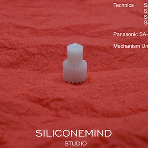 May include: A small, white plastic gear with a cylindrical top and toothed base, set against a textured red background. The image includes text indicating compatibility with Technics and Panasonic models, and the text "SILICONEMIND STUDIO".