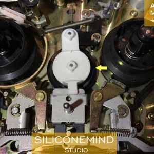 May include: Close-up of the inner workings of an Akai MG1212 cassette deck. The image shows a white plastic roller, a black metal reel, and a gold-colored metal lever.