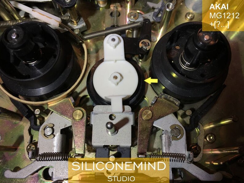 May include: Close-up of the inner workings of an Akai MG1212 cassette deck. The image shows a white plastic roller, a black metal reel, and a gold-colored metal lever.