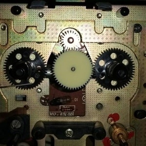 Puede incluir: Primer plano del mecanismo interno de un reproductor de casetes, que muestra una serie de engranajes y un engranaje de pl&aacute;stico blanco. La placa de metal est&aacute; marcada con el texto "674.001 - 202".