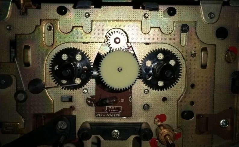 Puede incluir: Primer plano del mecanismo interno de un reproductor de casetes, que muestra una serie de engranajes y un engranaje de pl&aacute;stico blanco. La placa de metal est&aacute; marcada con el texto "674.001 - 202".