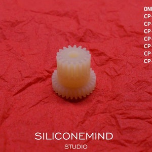 May include: A white plastic gear with a central hole. The gear is on a red background. The text "ONKYO" and a list of model numbers are visible in the background.