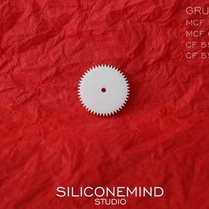 Puede incluir: Engranaje de pl&aacute;stico blanco con un agujero central. El engranaje est&aacute; sobre una superficie roja texturizada. El texto "GRUNDIG" es visible en la esquina superior derecha de la imagen. El texto "SILICONEMIND STUDIO" es visible en el centro inferior de la imagen.