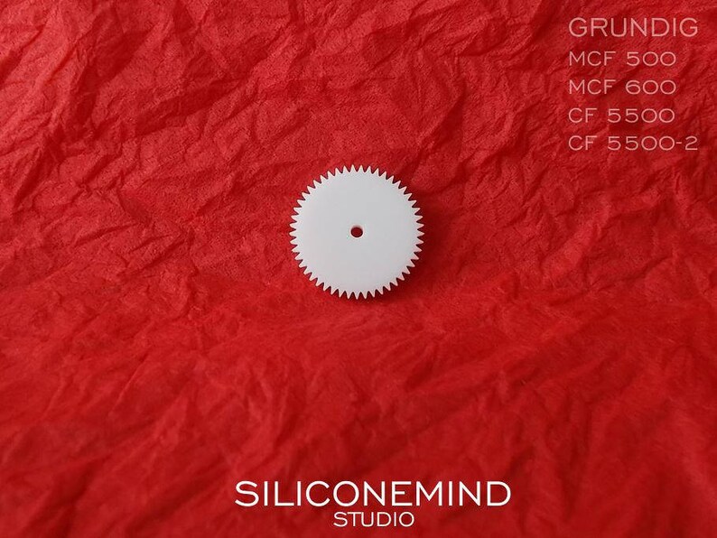 Puede incluir: Engranaje de pl&aacute;stico blanco con un agujero central. El engranaje est&aacute; sobre una superficie roja texturizada. El texto "GRUNDIG" es visible en la esquina superior derecha de la imagen. El texto "SILICONEMIND STUDIO" es visible en el centro inferior de la imagen.