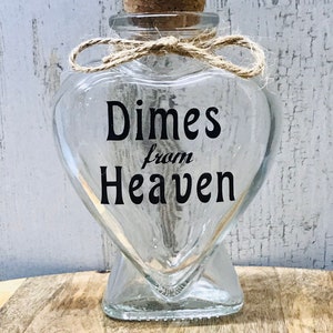 May include: A clear glass heart-shaped jar with a cork stopper and a twine bow. The jar has the text "Dimes from Heaven" printed on it.