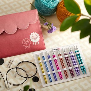 May include: A pink felt knitting needle case with a scalloped edge and sheep graphic. A set of colourful knitting needles in a white holder, yarn, and knitting accessories are also visible.