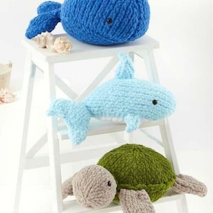 May include: A stack of three handmade crochet plush toys: a blue whale, a light blue dolphin, and a green turtle. The toys are displayed on a white step stool. The text "Jumbo Toastie" is visible.