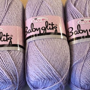 May include: Five skeins of lavender Babyglitz yarn. Each skein is wrapped with a pink label that reads "Babyglitz" in black script. The yarn has a subtle shimmer, suitable for baby projects. The yarn is a soft, light purple colour.