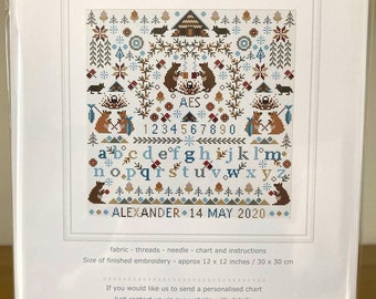 May include: A counted cross stitch sampler kit featuring a cabin in the woods with two bears, a fox, and a squirrel. The design includes the alphabet, numbers, and the text "Alexander 14 May 2020". The kit is 12 inches by 12 inches.