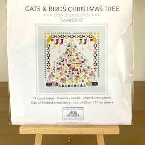 May include: A cross-stitch sampler kit titled "CATS & BIRDS CHRISTMAS TREE." The design features a Christmas tree decorated with cats, birds, and ornaments. The kit includes 14-count fabric, threads, a needle, and instructions. Finished embroidery size is approximately 10 inches square.
