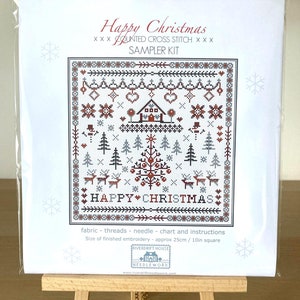 May include: A white cross stitch sampler kit with a winter scene featuring a house, trees, reindeer, and the text "Happy Christmas". The kit includes fabric, threads, a needle, a chart, and instructions. The finished embroidery will be approximately 10 inches square.
