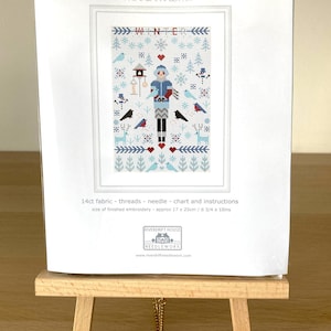 May include: A white card with the text "MINI WINTER SAMPLER" and "X X X COUNTED CROSS STITCH X X X" and "MINI SAMPLER KIT". The card features a cross stitch pattern of a winter scene with a person in a blue coat and hat, birds, trees, and snowflakes. The card is on a small wooden easel. The text "14ct fabric - threads - needle - chart and instructions" and "size of finished embroidery - approx 17 x 25cm / 6 3/4 x 10ins" is also on the card.