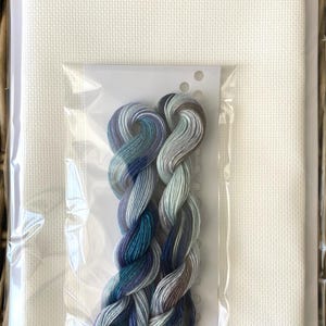 May include: A cross-stitch kit featuring white fabric and a skein of colorful embroidery floss. The floss is a blend of blues, greens, and gray. The kit is packaged in a clear plastic bag, ready for a crafting project.