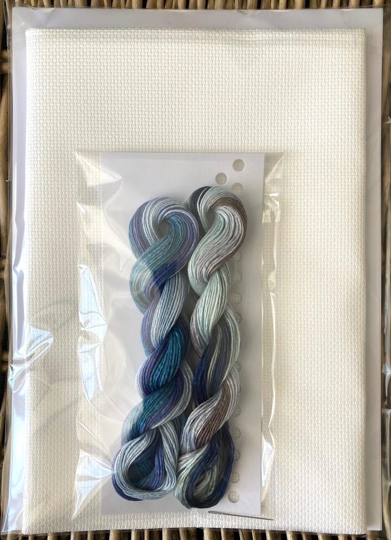 May include: A cross-stitch kit featuring white fabric and a skein of colorful embroidery floss. The floss is a blend of blues, greens, and gray. The kit is packaged in a clear plastic bag, ready for a crafting project.