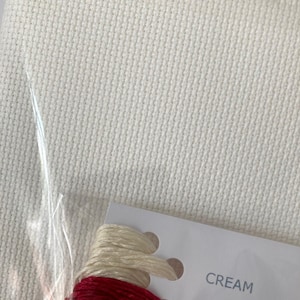 Pu&ograve; includere: Primo piano di un tessuto Aida bianco con una carta che mostra due gomitoli di filo da ricamo. La carta dice "CREAM" e "CHERRY RED".