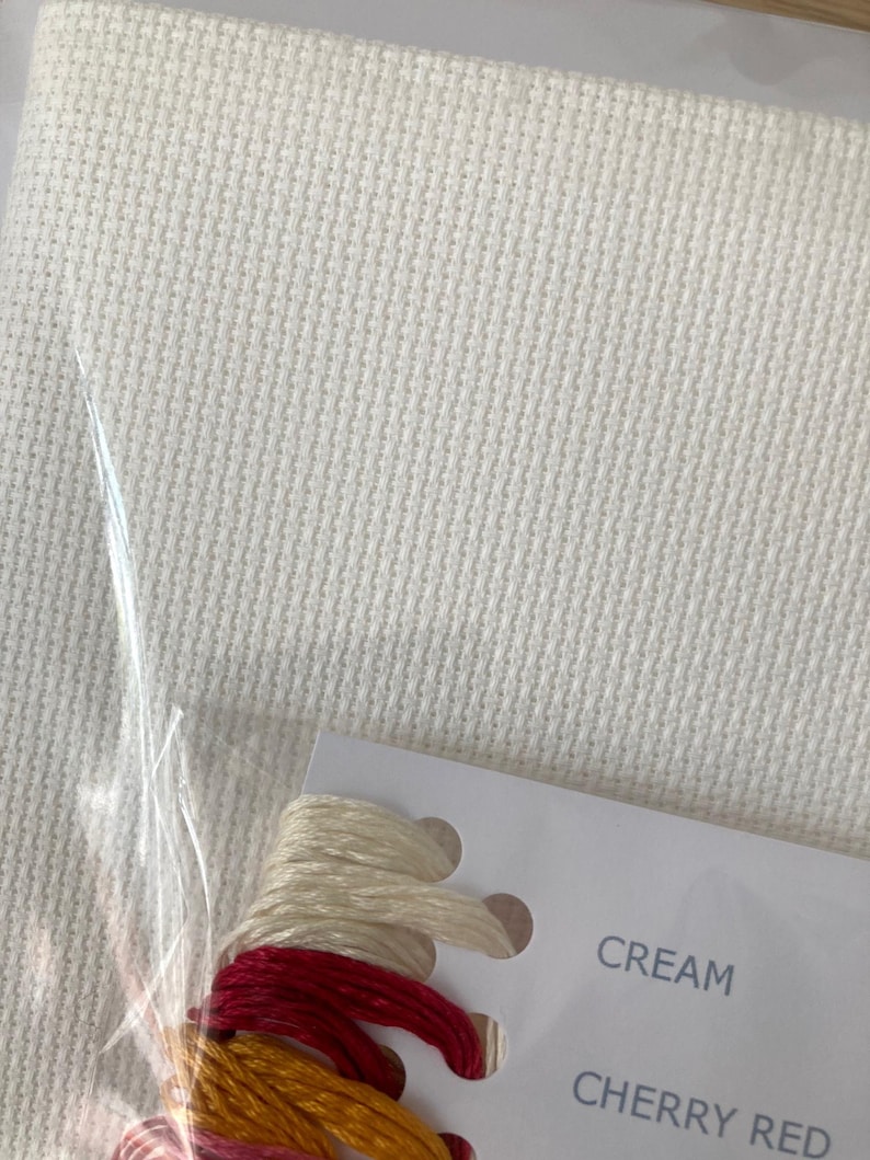 Pu&ograve; includere: Primo piano di un tessuto Aida bianco con una carta che mostra due gomitoli di filo da ricamo. La carta dice "CREAM" e "CHERRY RED".