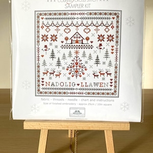 May include: A cross stitch sampler kit with a winter scene featuring a house, a Christmas tree, and reindeer. The kit includes fabric, threads, a needle, a chart, and instructions. The finished embroidery will be approximately 10 inches square.