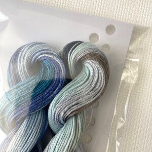 May include: Close-up of embroidery floss in shades of blue, teal, gray, and brown. The floss is twisted and bundled, partially visible through a clear plastic package. The background is a white, textured fabric.