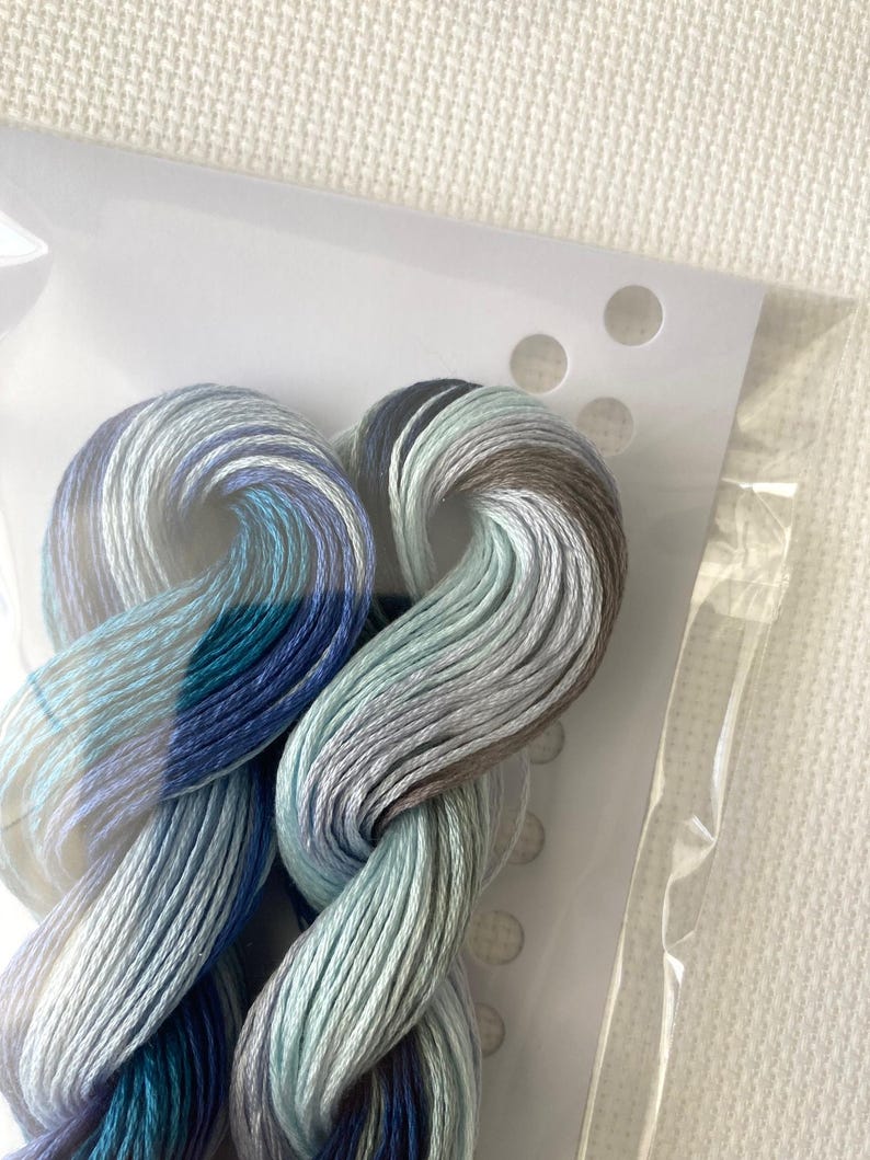 May include: Close-up of embroidery floss in shades of blue, teal, gray, and brown. The floss is twisted and bundled, partially visible through a clear plastic package. The background is a white, textured fabric.