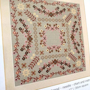 May include: A counted cross stitch kit featuring a floral design. The embroidery pattern includes a square border with flowers and leaves in shades of brown, pink, and beige on a gray background. The finished embroidery size is approximately 12 inches square.