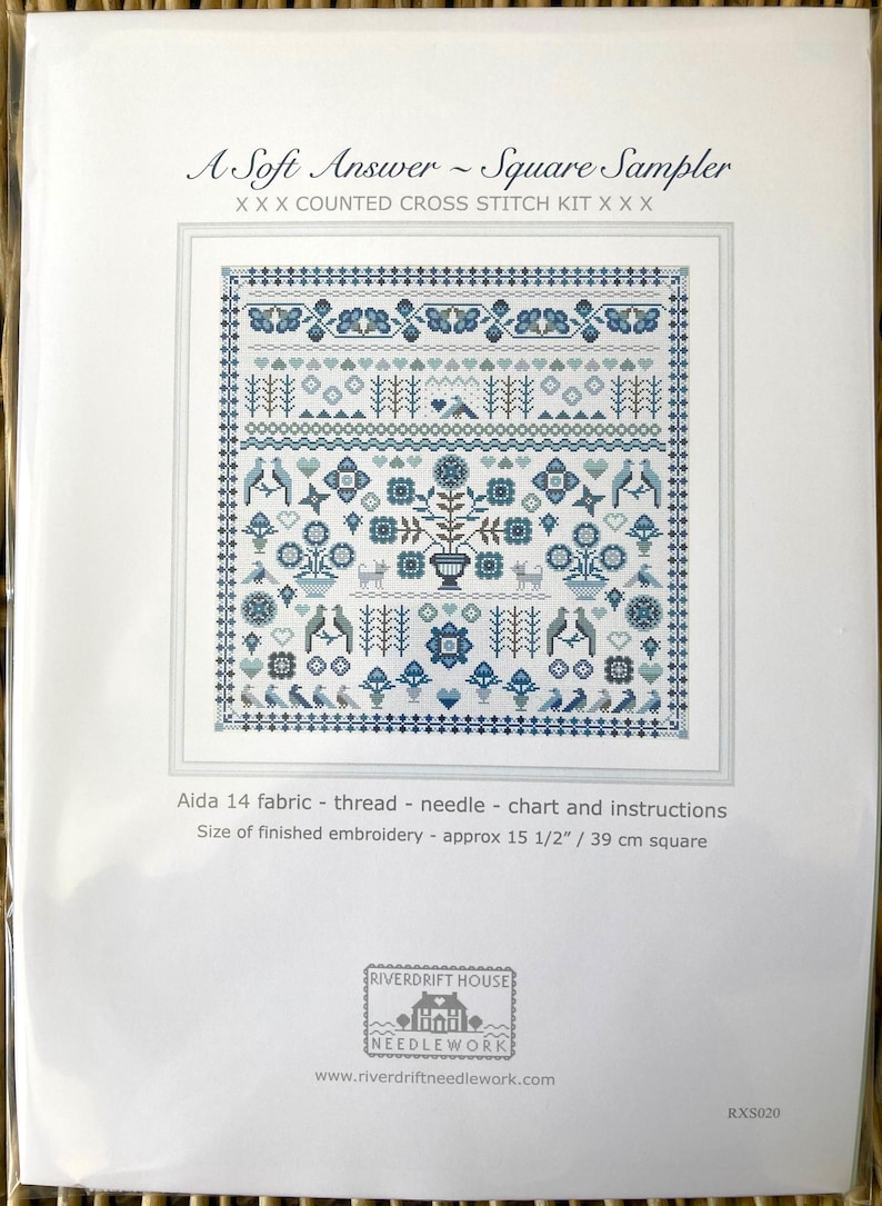 May include: A counted cross stitch kit titled "A Soft Answer - Square Sampler." The design features a detailed, symmetrical pattern in shades of blue and white, including birds, flowers, and geometric shapes. The finished embroidery size is approximately 15 1/2 inches square.