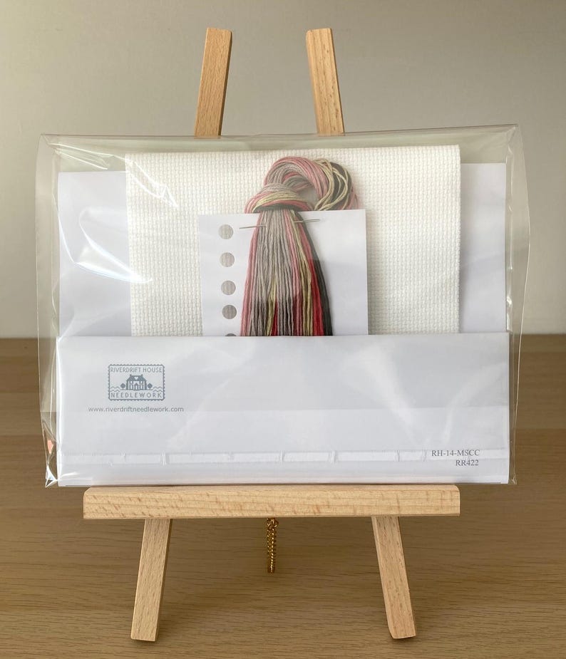 May include: A cross stitch kit with white Aida fabric, floss, and a needle. The kit is packaged in a clear plastic bag and is displayed on a wooden easel. The kit includes the text "Riverdrift House Needlework" and "RHF-14-MSCC RR422".