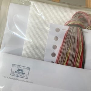 May include: A cross stitch kit with white Aida cloth, floss in shades of pink, beige, and green, a needle, and a small card with the text "Riverdrift House Needlework" and a website address.