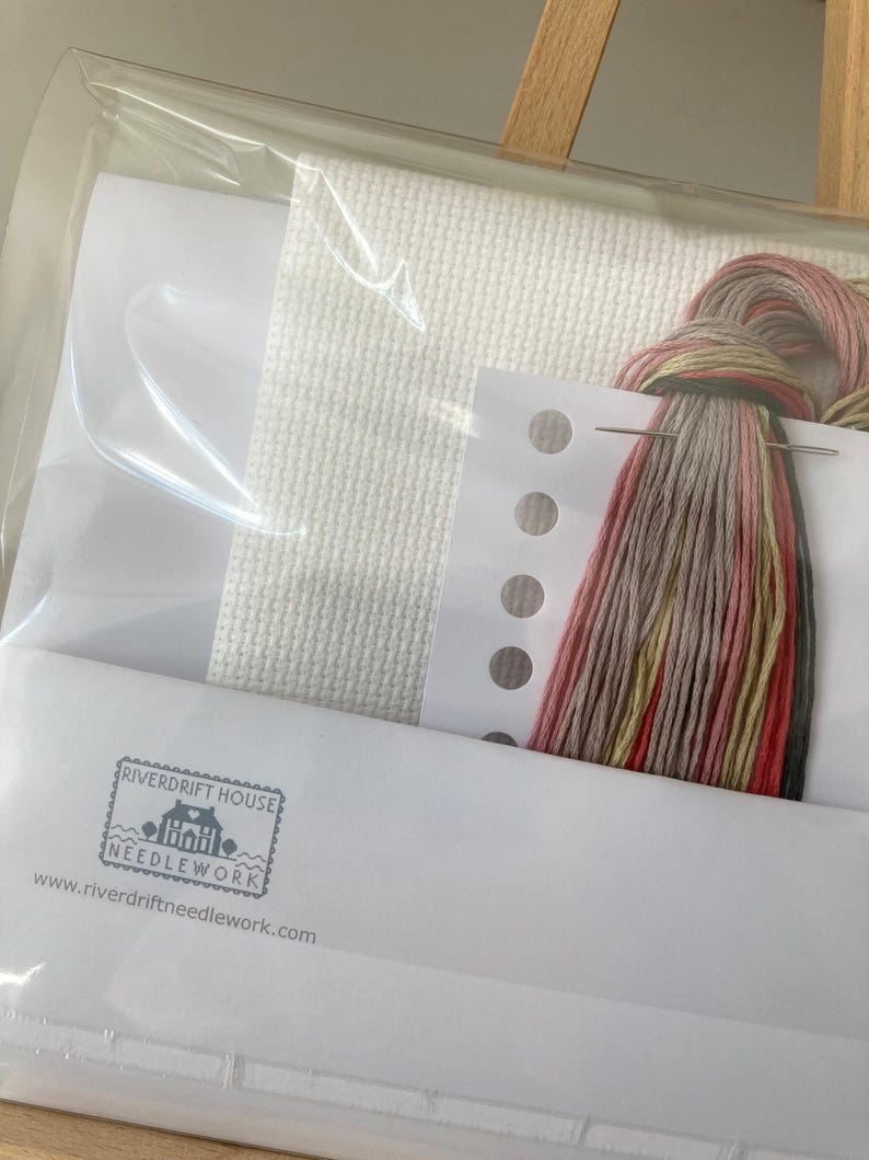 May include: A cross stitch kit with white Aida cloth, floss in shades of pink, beige, and green, a needle, and a small card with the text "Riverdrift House Needlework" and a website address.