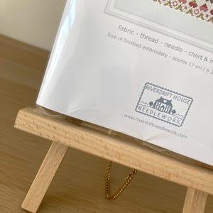 May include: A white card with a cross-stitch pattern on it. The card is on a wooden easel with a gold chain. The card has the text "fabric - thread - needle - chart & in" and "Size of finished embroidery - approx 17 cm / 6.3" printed on it. The card also has a stamp with the text "RIVERDRIFT HOUSE NEEDLEWORK" and the website address "www.riverdriftneedlework.com".