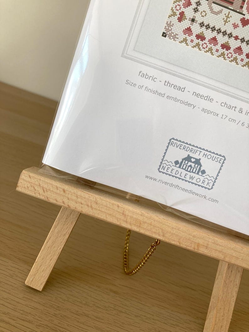 May include: A white card with a cross-stitch pattern on it. The card is on a wooden easel with a gold chain. The card has the text "fabric - thread - needle - chart & in" and "Size of finished embroidery - approx 17 cm / 6.3" printed on it. The card also has a stamp with the text "RIVERDRIFT HOUSE NEEDLEWORK" and the website address "www.riverdriftneedlework.com".