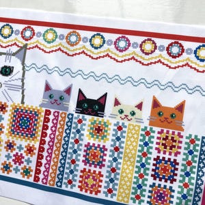 May include: White fabric with a cross-stitch design. It features a large gray cat and four smaller cats. The design includes colourful geometric patterns, a red border, and a wavy blue line.