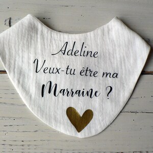 May include: White fabric bib with black text that reads "Adeline Veux-tu &ecirc;tre ma Marraine?" and a gold heart.