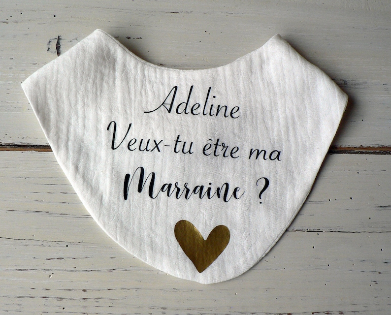 May include: White fabric bib with black text that reads "Adeline Veux-tu &ecirc;tre ma Marraine?" and a gold heart.