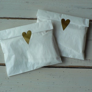 May include: Two white paper bags with gold heart stickers on the front. The bags are sitting on a white wooden surface.