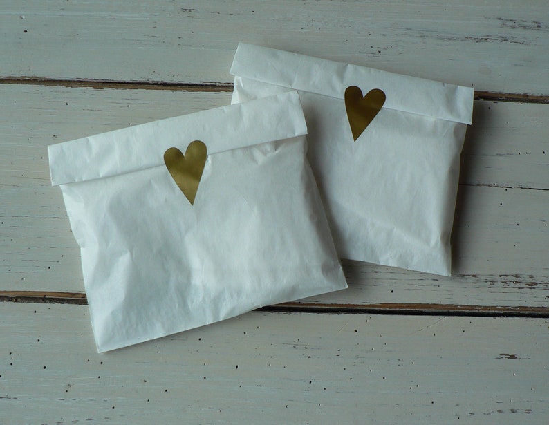 May include: Two white paper bags with gold heart stickers on the front. The bags are sitting on a white wooden surface.
