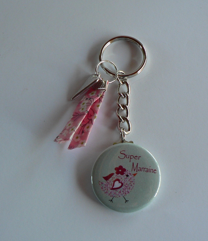 Puede incluir: Un llavero con un dije redondo azul claro con un dise&ntilde;o de p&aacute;jaro y el texto "Super Marraine" en rosa. El llavero incluye una cadena plateada, un llavero y cintas florales rosas.